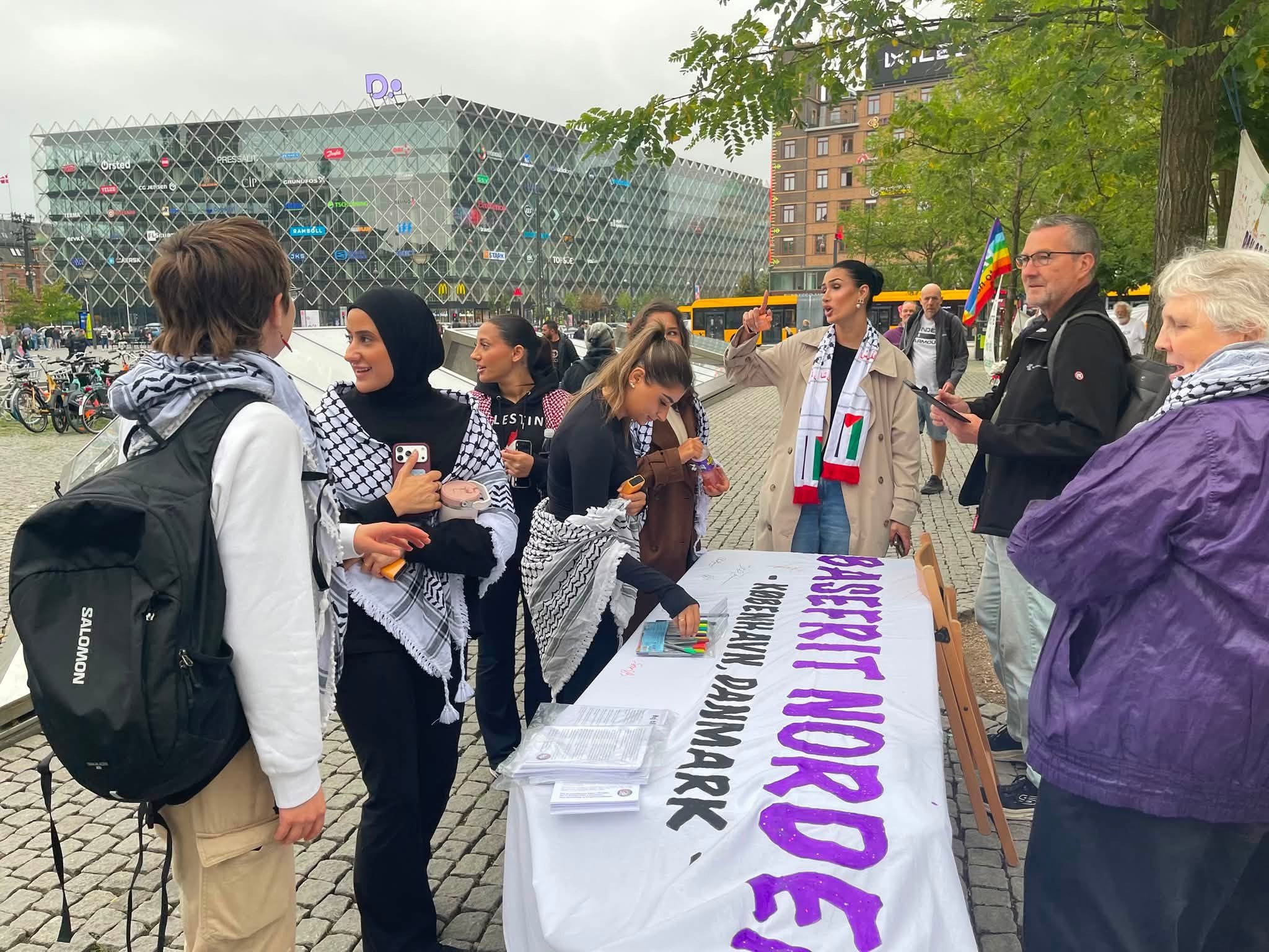 Aktivister og besøkende ved et bord med løpesedler og banneret «Basefrit Norden» og «København, Danmark».