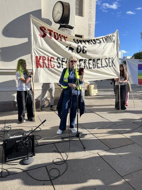En kvinne holder appell ved en mikrofon foran et banner mot opprustning og krigsforberedelser i Oslo.