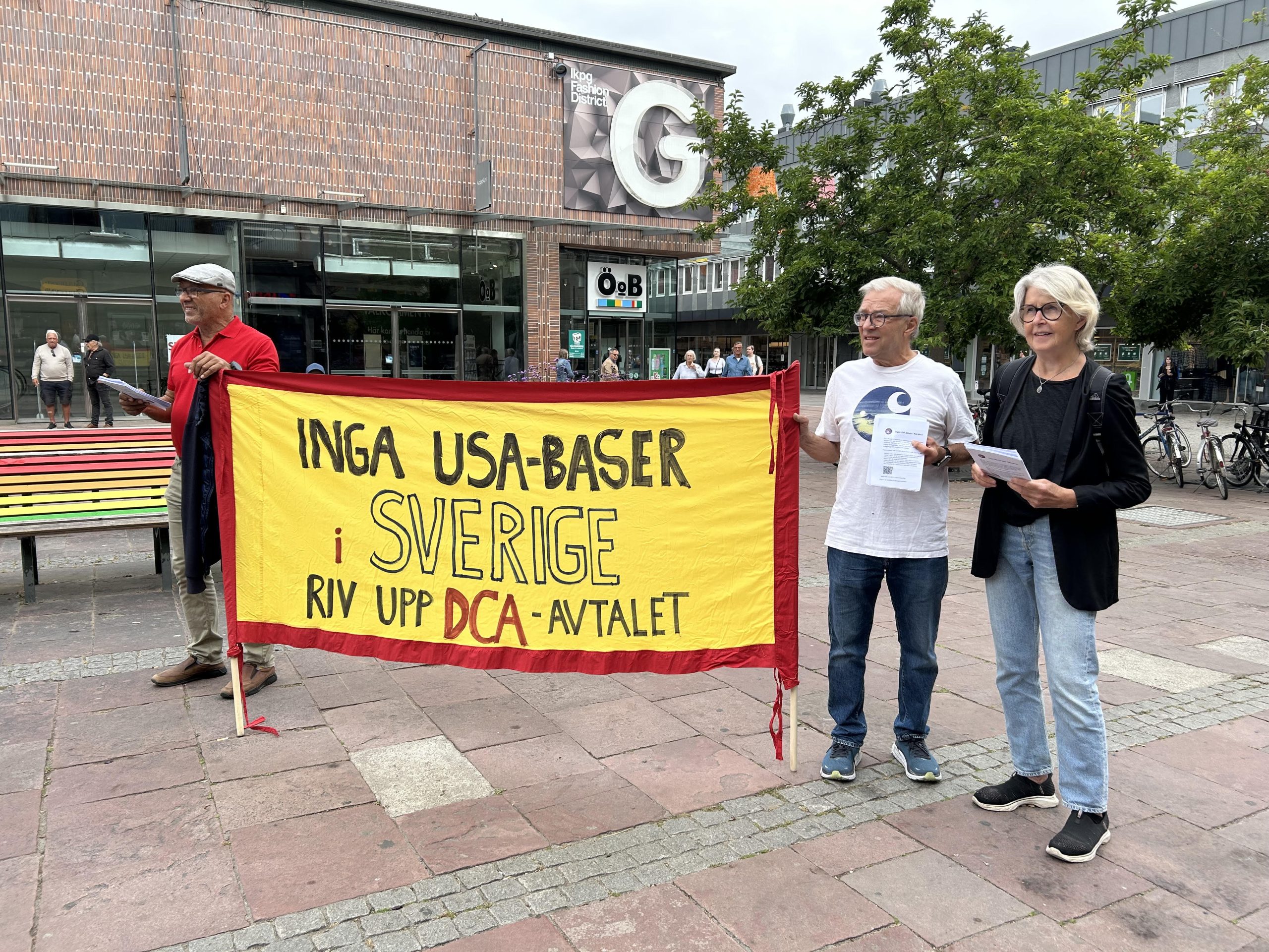 Tre aktivister holder et gult banner med teksten «Inga USA-baser i Sverige – riv upp DCA-avtalet».